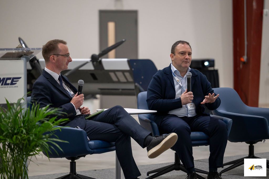 Two men in suits sit on blue conference chairs on a stage, each holding a microphone and talking during a panel discussion, with a small table between them and a display aircraft in the background.