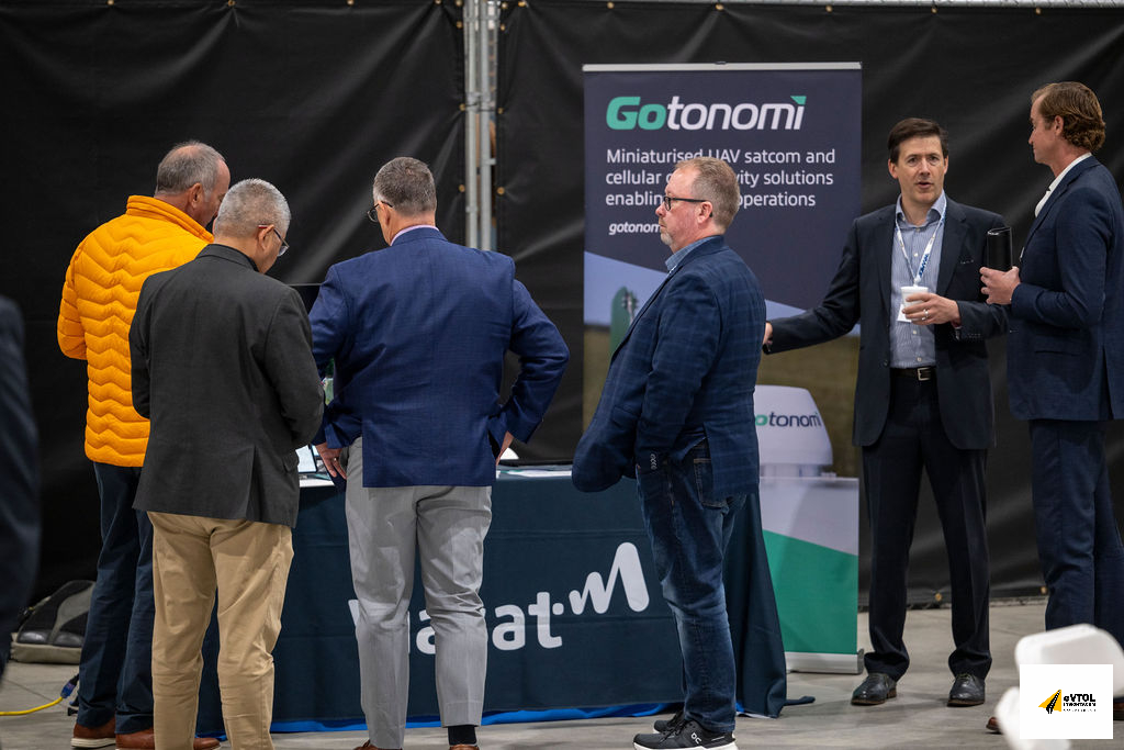 Five men in business attire stand around a conference booth; one wears an orange puffer jacket while others chat near a Gotonomi banner.