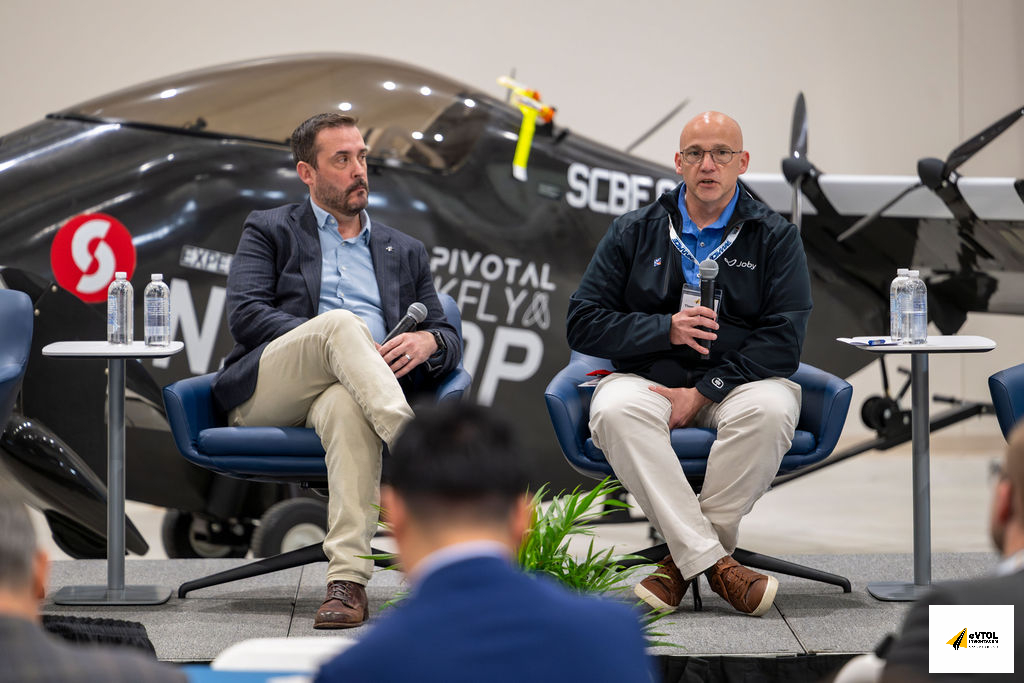 Two men sit on stage in blue chairs, each holding a microphone, with a small airplane in the background at a panel discussion in-flight expo setting.