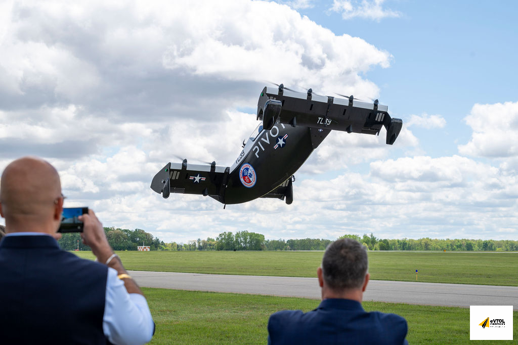 Tilt-rotor military aircraft lifting off over an airfield, two spectators filming from the foreground.