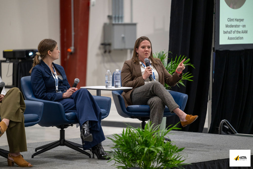 Two female panelists on a stage, one speaking into a microphone, the other listening, with a screen and plants nearby