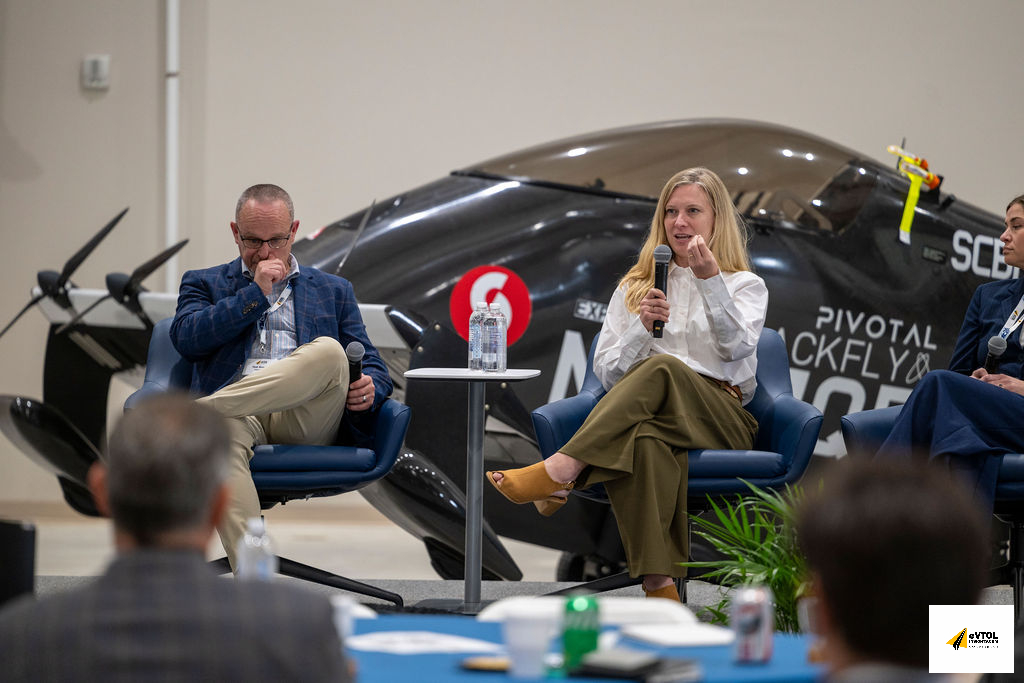 Panel discussion in a hangar with a sleek black airplane in the background; three speakers sit in blue chairs, the center woman speaks into a microphone while gesturing.