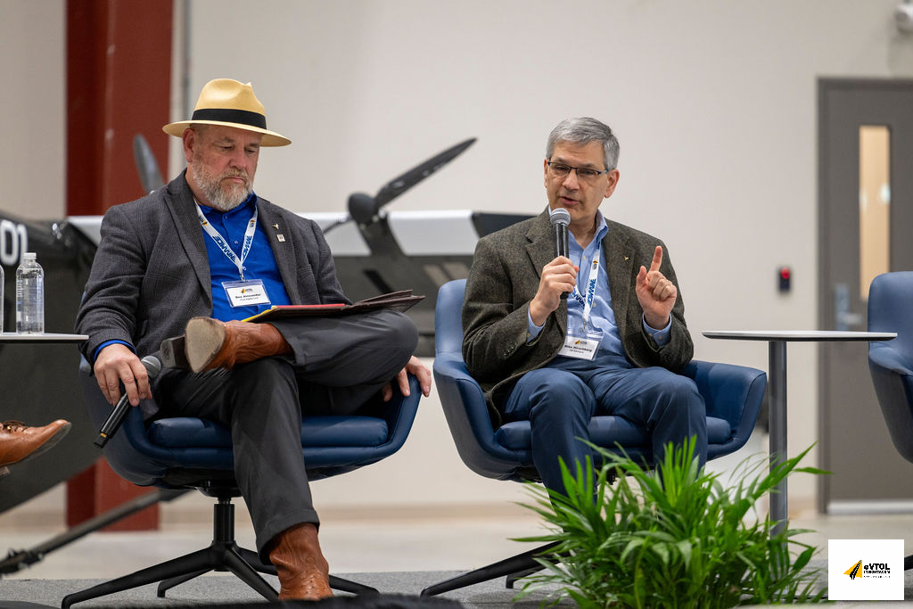 Panel discussion: man on the right with a microphone speaking, the man on the left looking over notes; both wear blazers and name badges, seated on stage with a plant in front.