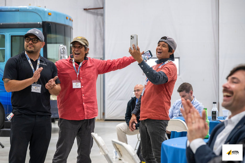 Three men with lanyards cheer and pose for a selfie indoors during an event and celebration.