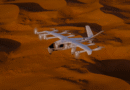 White tilt-rotor aircraft flying low over orange desert sand dunes.