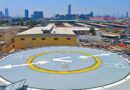 Rooftop helicopter pad with a large V and arrows, yellow ring, and surrounding industrial buildings against a clear blue sky in a cityscape.