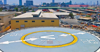 Rooftop helicopter pad with a large V and arrows, yellow ring, and surrounding industrial buildings against a clear blue sky in a cityscape.