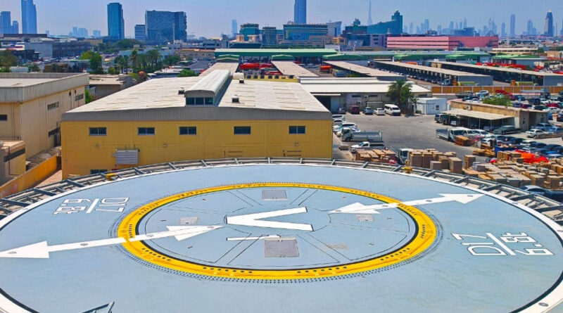 Rooftop helicopter pad with a large V and arrows, yellow ring, and surrounding industrial buildings against a clear blue sky in a cityscape.