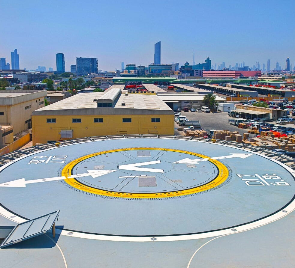Rooftop helicopter pad with a large V and arrows, yellow ring, and surrounding industrial buildings against a clear blue sky in a cityscape.