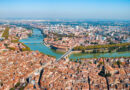Aerial cityscape with a winding river, multiple bridges, and dense red-roofed buildings along green riverbanks.