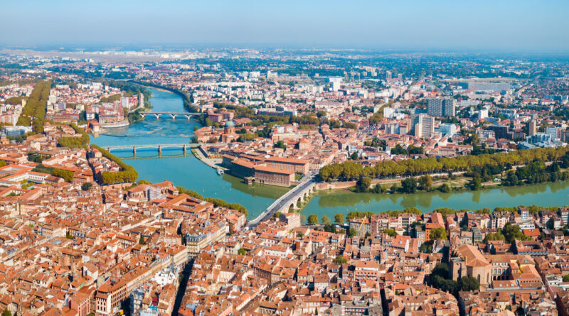 Aerial cityscape with a winding river, multiple bridges, and dense red-roofed buildings along green riverbanks.