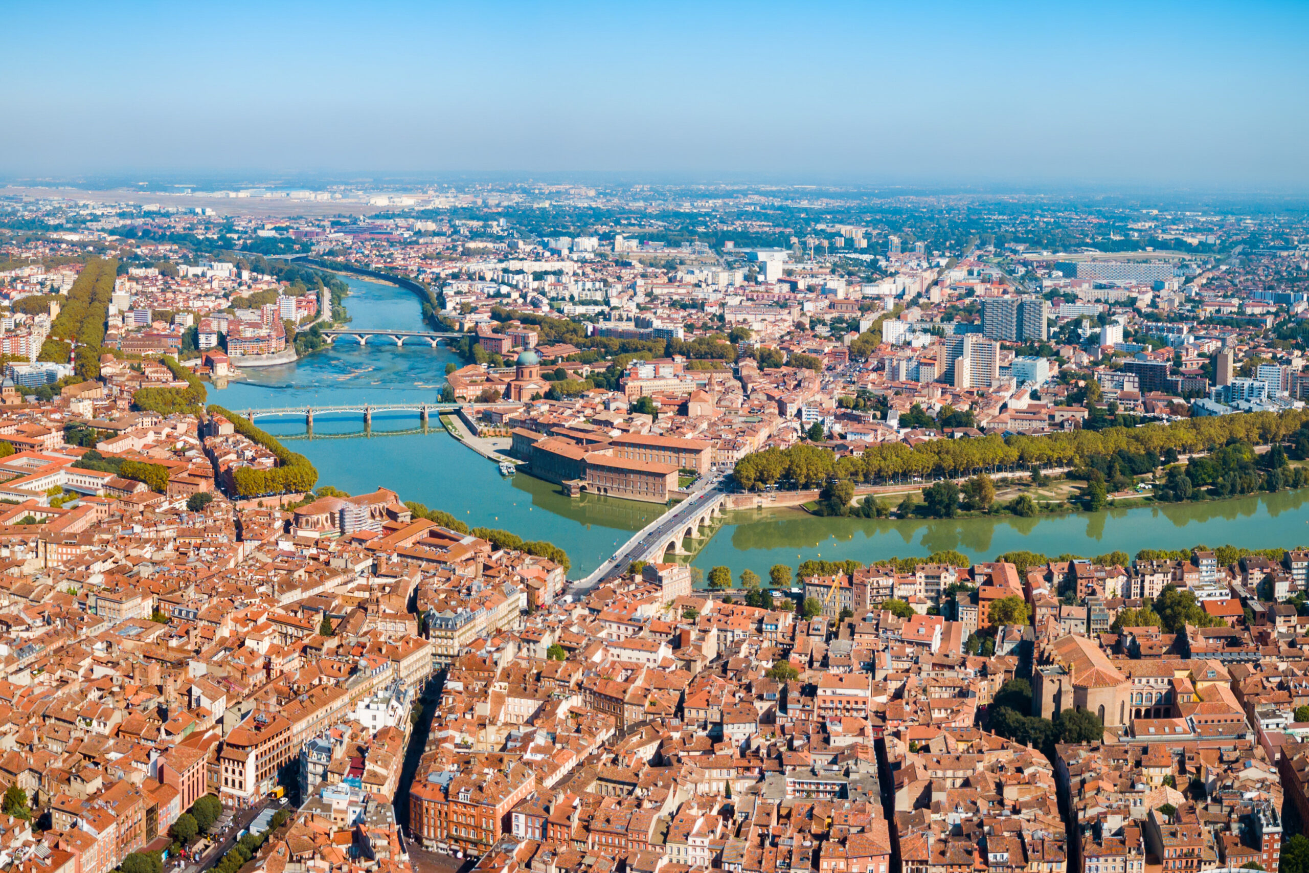 Aerial cityscape with a winding river, multiple bridges, and dense red-roofed buildings along green riverbanks.