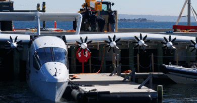 image001 10 - eVTOL Insights White seaplane moored at a wooden dock, with propeller engines along the wing; crew members stand on the deck behind a railing, a red life ring hangs from the side, and calm sea with distant coastline is in the background.