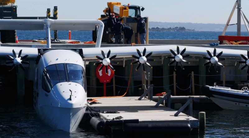 image001 10 - eVTOL Insights White seaplane moored at a wooden dock, with propeller engines along the wing; crew members stand on the deck behind a railing, a red life ring hangs from the side, and calm sea with distant coastline is in the background.