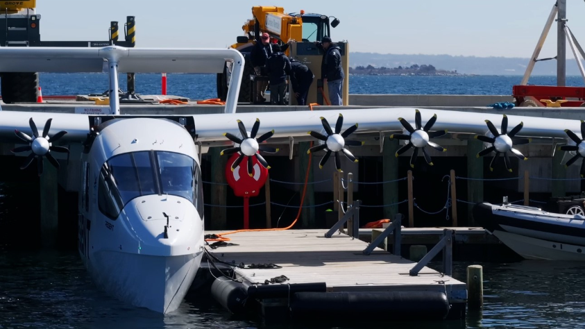 White seaplane moored at a wooden dock, with propeller engines along the wing; crew members stand on the deck behind a railing, a red life ring hangs from the side, and calm sea with distant coastline is in the background.