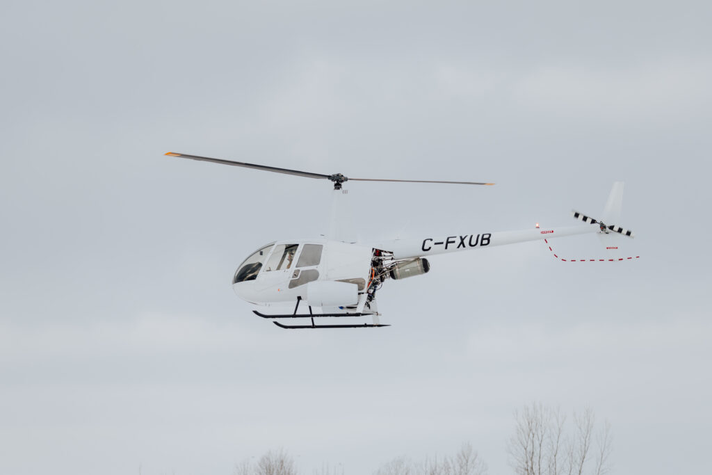 White helicopter in flight against a pale, overcast sky with rotor blades spinning and landing skids visible beneath the fuselage (registration C-FXUB).