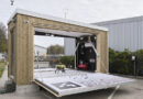 Mobile wooden lab with a wide fold-down ramp; inside a small drone sits on a platform beside equipment like a compressor/generator. The unit is outdoors near a gray industrial building.