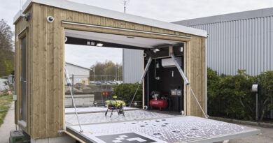 Mobile wooden lab with a wide fold-down ramp; inside a small drone sits on a platform beside equipment like a compressor/generator. The unit is outdoors near a gray industrial building.