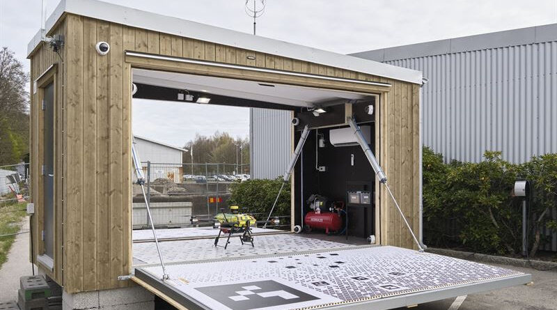 Mobile wooden lab with a wide fold-down ramp; inside a small drone sits on a platform beside equipment like a compressor/generator. The unit is outdoors near a gray industrial building.