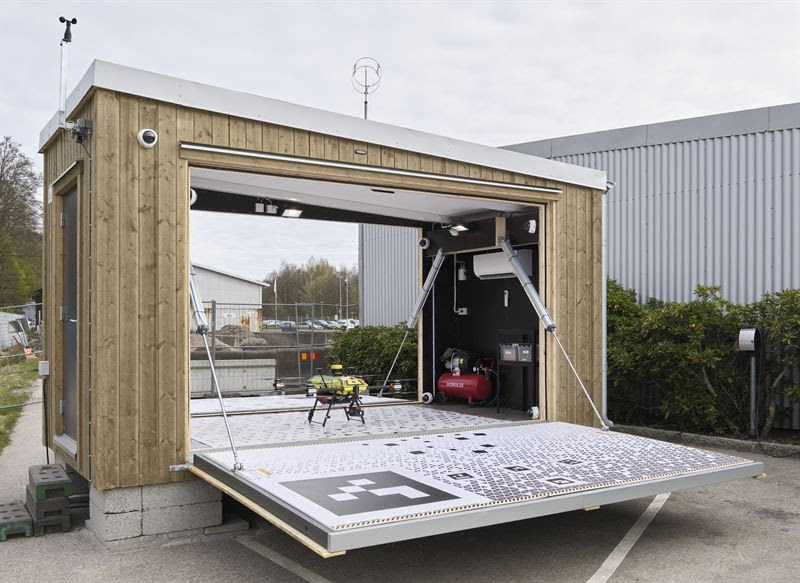 Mobile wooden lab with a wide fold-down ramp; inside a small drone sits on a platform beside equipment like a compressor/generator. The unit is outdoors near a gray industrial building.