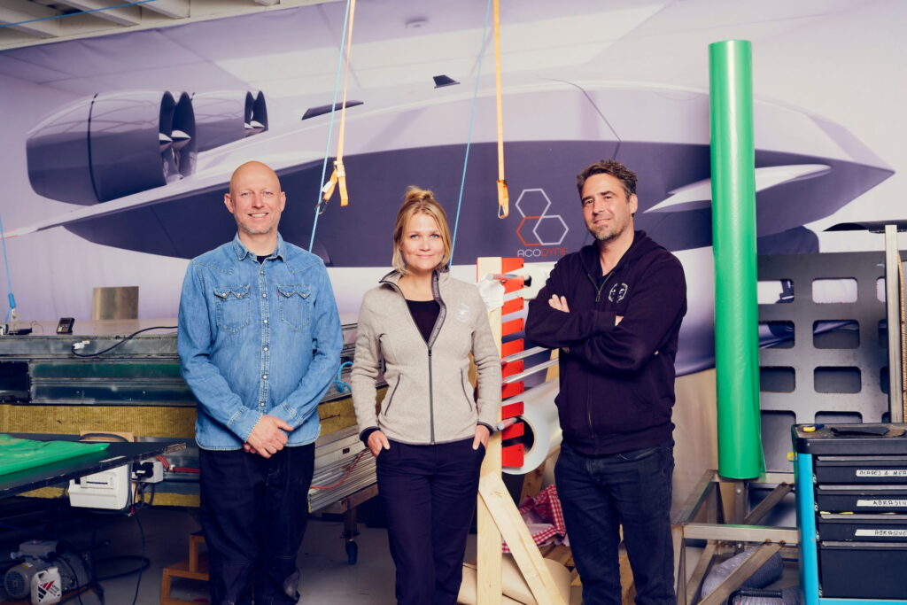 04-Acodyne-_DSF3177 1 - eVTOL Insights Three workers pose in a hangar-style workshop in front of a large airplane mural; left man in a denim shirt, center woman in a gray zip jacket, right man in a black hoodie