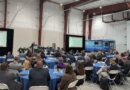 1000032269 - eVTOL Insights Large conference in a hangar with round tables, attendees in suits, and two presentation screens show slides; a blue SkyVision bus is parked in the background.