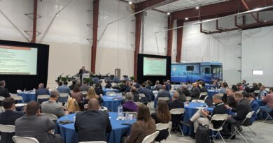 Large conference in a hangar with round tables, attendees in suits, and two presentation screens show slides; a blue SkyVision bus is parked in the background.