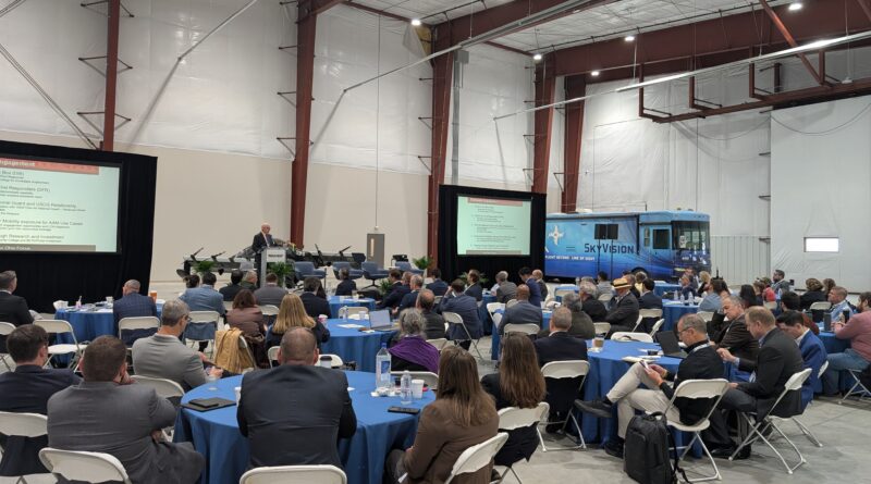 Large conference in a hangar with round tables, attendees in suits, and two presentation screens show slides; a blue SkyVision bus is parked in the background.