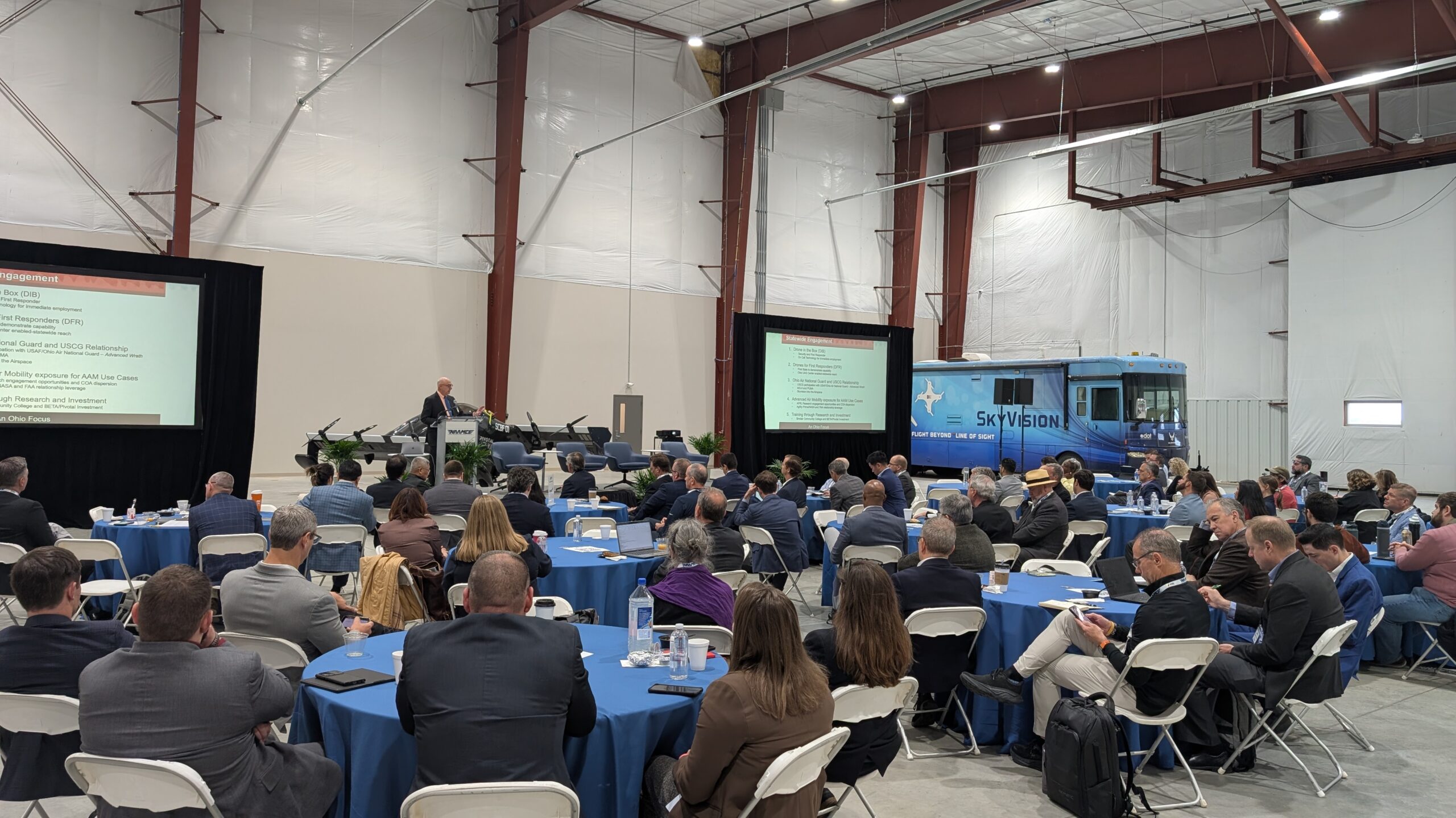 Large conference in a hangar with round tables, attendees in suits, and two presentation screens show slides; a blue SkyVision bus is parked in the background.