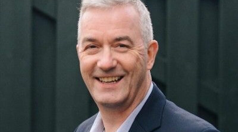 Smiling man with short gray hair in a blazer and light shirt, outdoors against a dark fence backdrop.