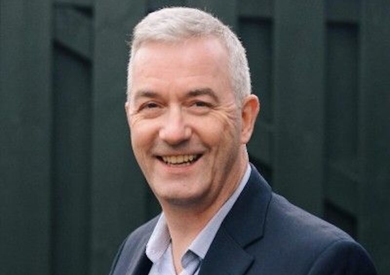 Smiling man with short gray hair in a blazer and light shirt, outdoors against a dark fence backdrop.