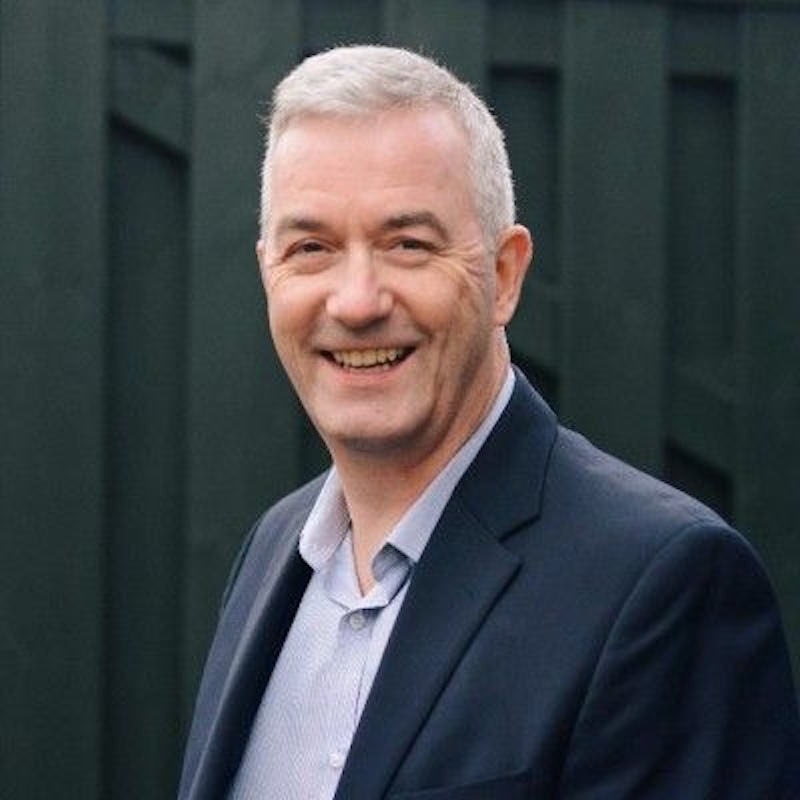 Smiling man with short gray hair in a blazer and light shirt, outdoors against a dark fence backdrop.