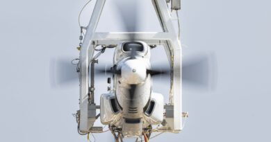 Front view of a white aircraft nose held in a gray structural frame with a spinning propeller in motion, against a pale sky backdrop.