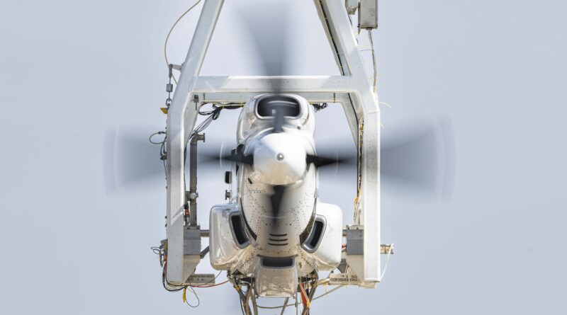 Front view of a white aircraft nose held in a gray structural frame with a spinning propeller in motion, against a pale sky backdrop.