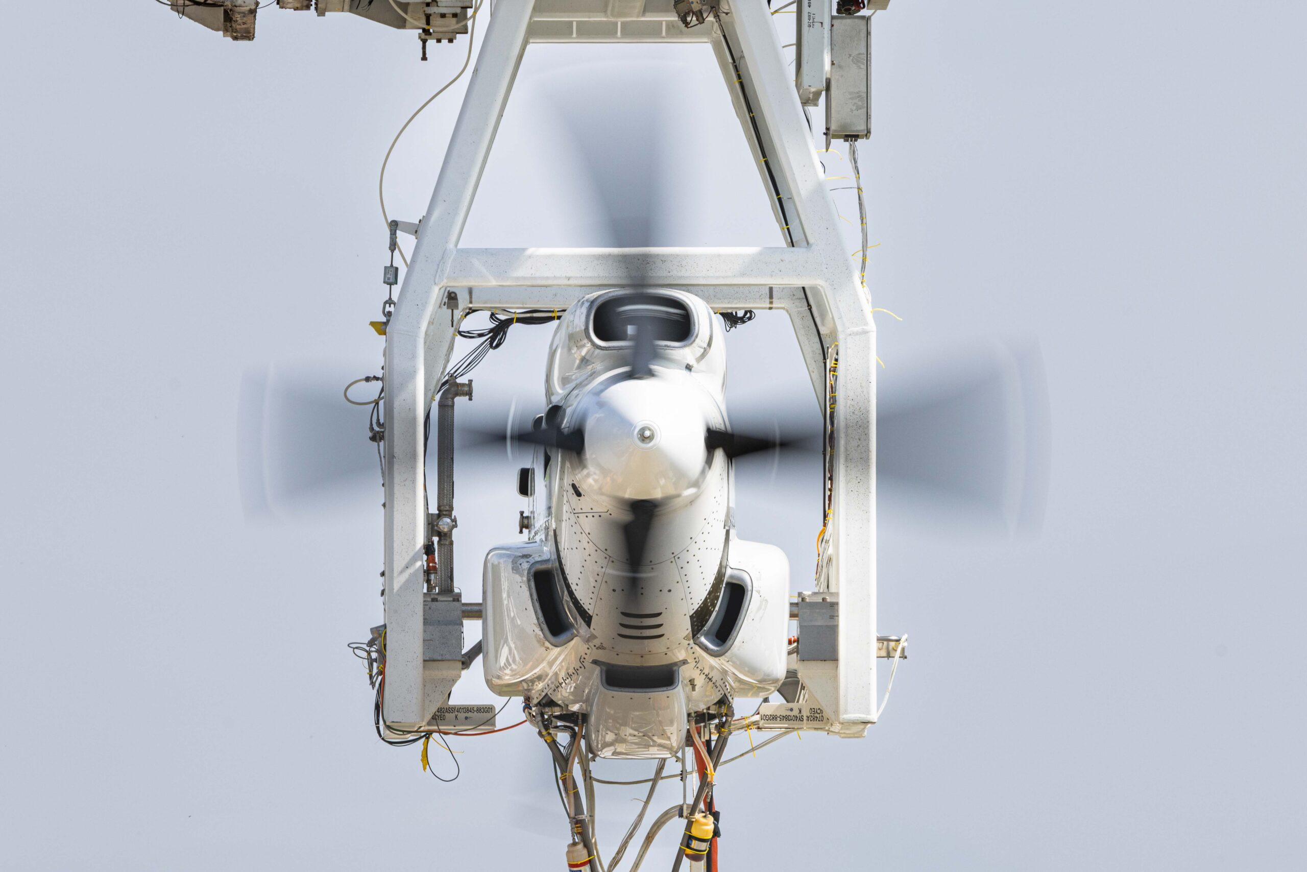 Front view of a white aircraft nose held in a gray structural frame with a spinning propeller in motion, against a pale sky backdrop.