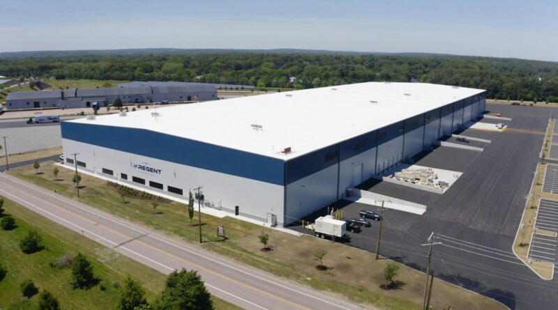 Aerial view of a large white-roofed warehouse with blue side walls, loading docks, and adjacent parking areas labeled Regent.
