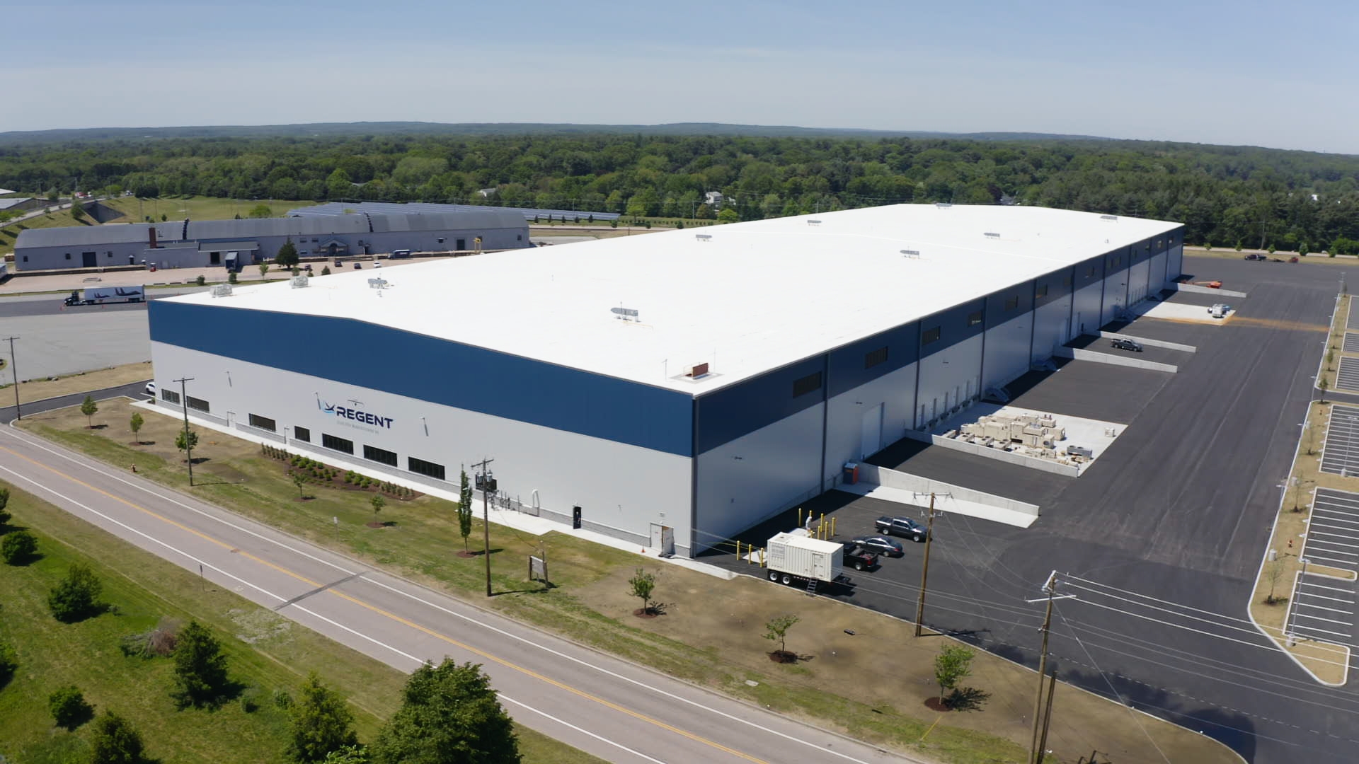 Aerial view of a large white-roofed warehouse with blue side walls, loading docks, and adjacent parking areas labeled Regent.