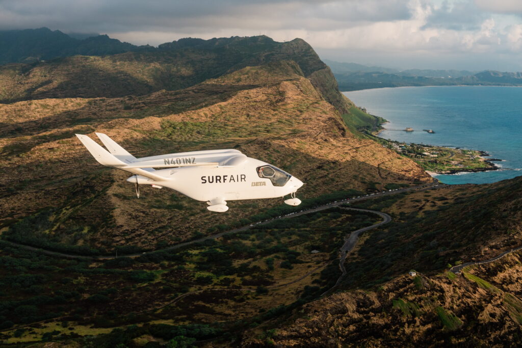 Small white SurfAir airplane with N401NZ visible, flying over rugged coastal hills as a winding road passes below and the blue ocean stretches along the shore to the right.