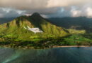 Small white airplane labeled SURFAIR flying over lush green tropical mountains and a coastal shoreline.
