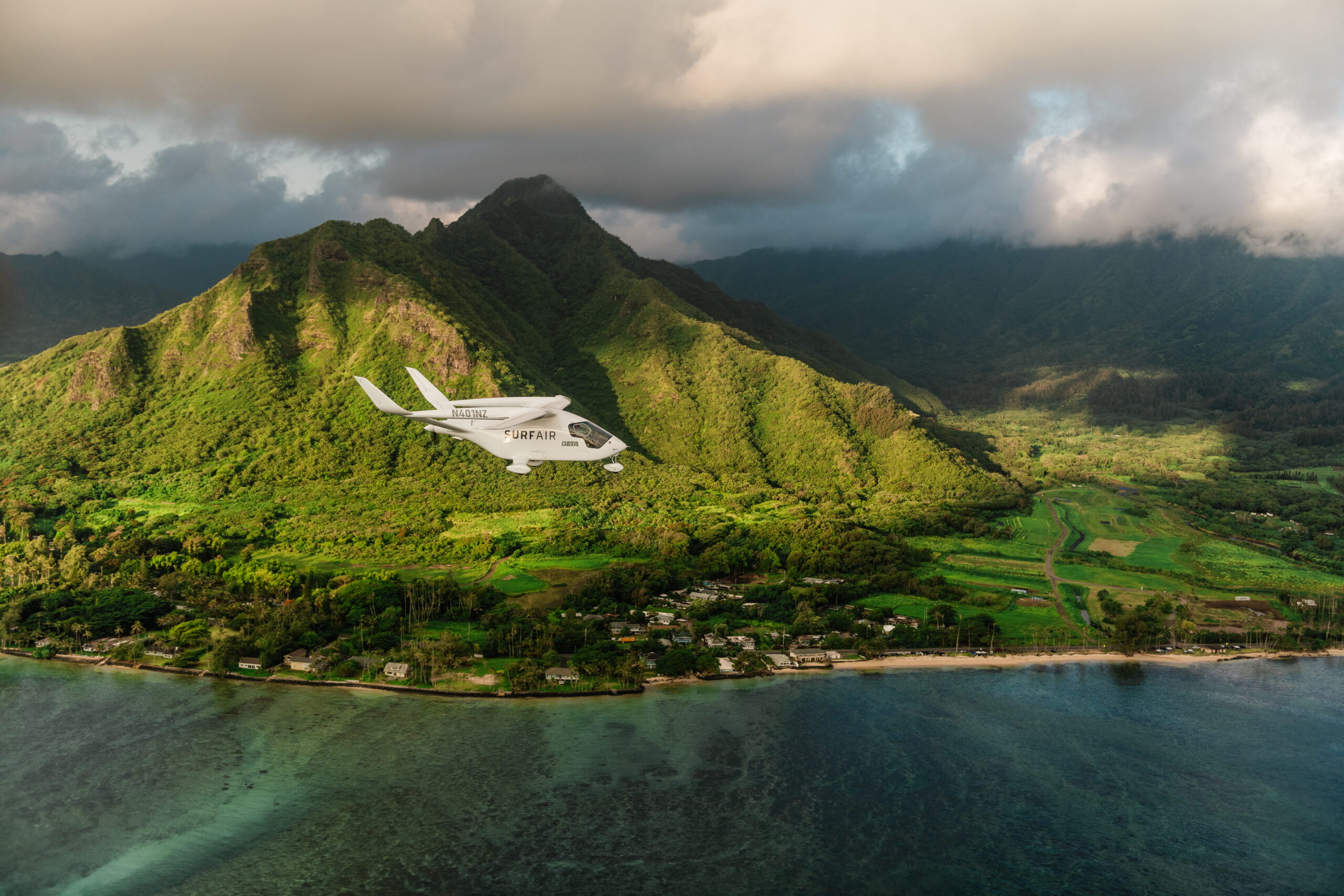 Small white airplane labeled SURFAIR flying over lush green tropical mountains and a coastal shoreline.