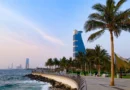 Seaside promenade with palm trees, a curved railing, and a blue futuristic skyscraper along the coast at sunset.