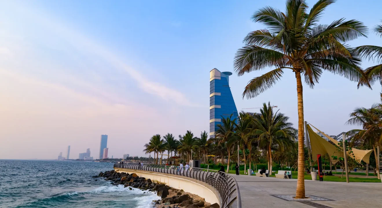 Seaside promenade with palm trees, a curved railing, and a blue futuristic skyscraper along the coast at sunset.
