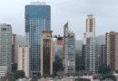 Drone carrying an iFood delivery box flies over a city skyline with tall buildings in the background.