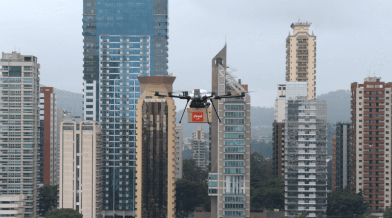 Drone carrying an iFood delivery box flies over a city skyline with tall buildings in the background.