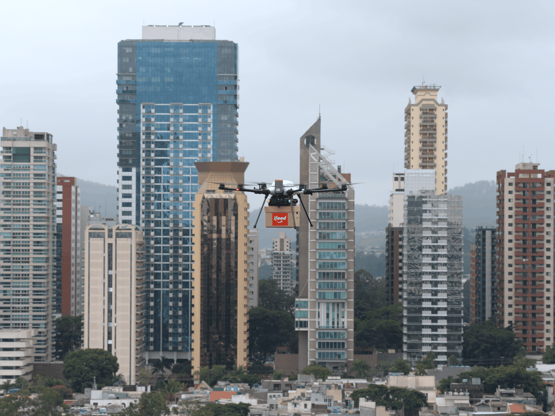 Drone carrying an iFood delivery box flies over a city skyline with tall buildings in the background.