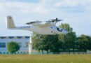 White tilt-rotor aircraft (V-22/Osprey style) hovering low above a grassy field, registration G-EVTB, with trees and a building in the background.