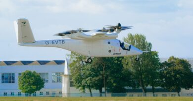 White tilt-rotor aircraft (V-22/Osprey style) hovering low above a grassy field, registration G-EVTB, with trees and a building in the background.