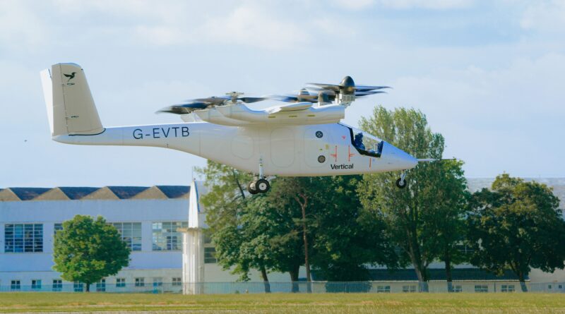White tilt-rotor aircraft (V-22/Osprey style) hovering low above a grassy field, registration G-EVTB, with trees and a building in the background.