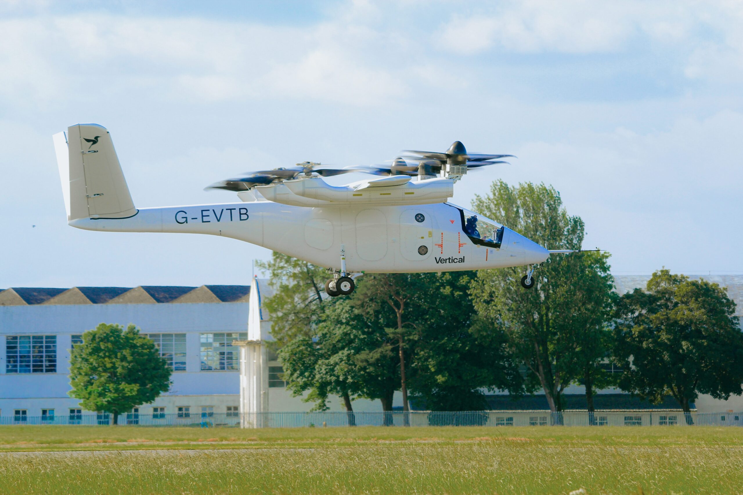 White tilt-rotor aircraft (V-22/Osprey style) hovering low above a grassy field, registration G-EVTB, with trees and a building in the background.