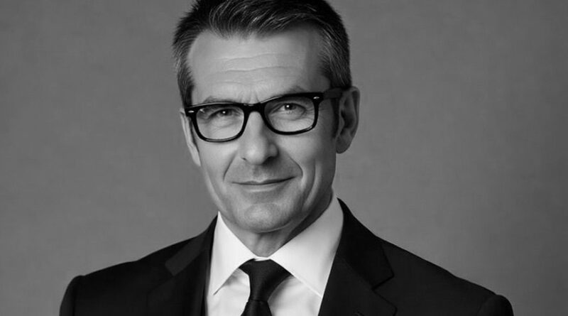 Portrait of a middle-aged man in a black suit, white shirt, black tie, and glasses looking at the camera.